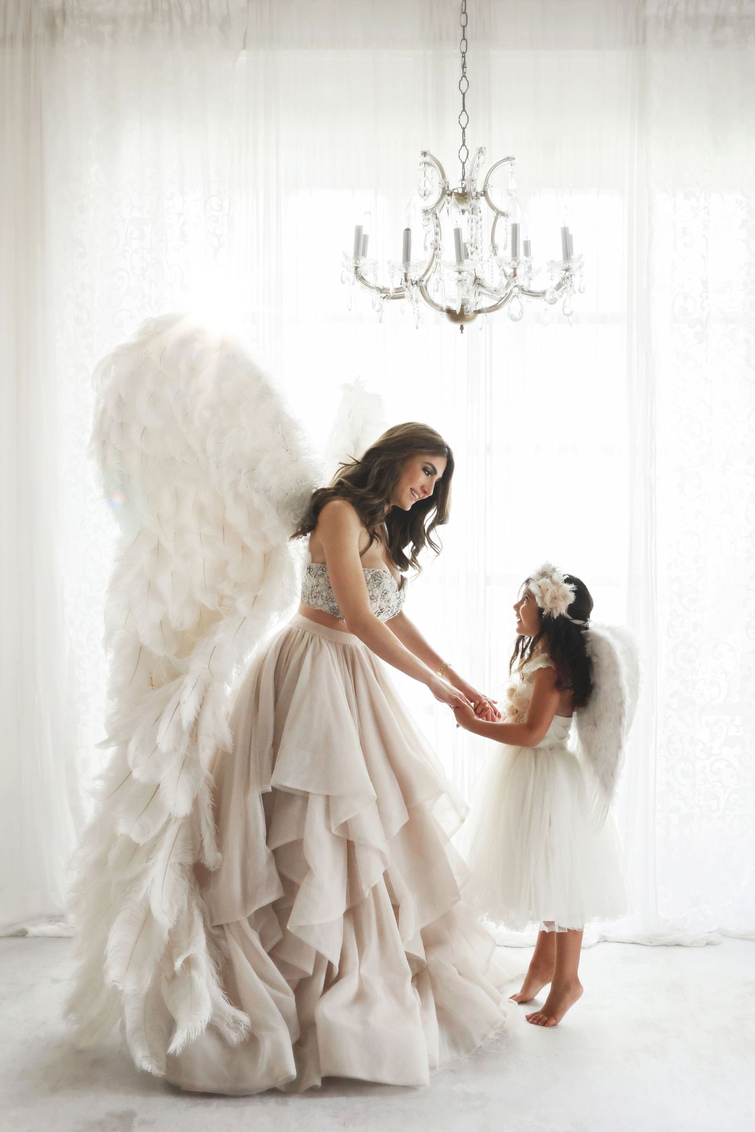Mother and daughter in angel wings holding hands in bright white studio with chandelier.