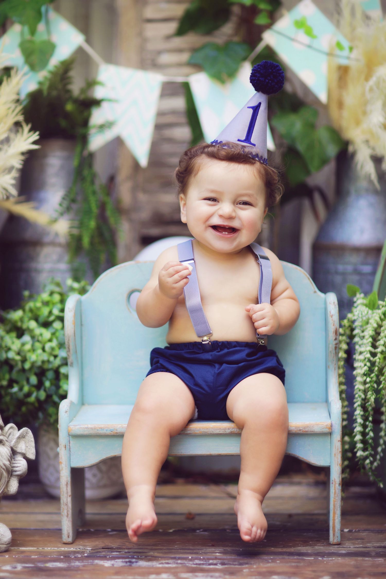 Smiling baby boy in navy shorts and suspenders celebrates his 1st birthday wearing a blue party hat on a rustic bench.