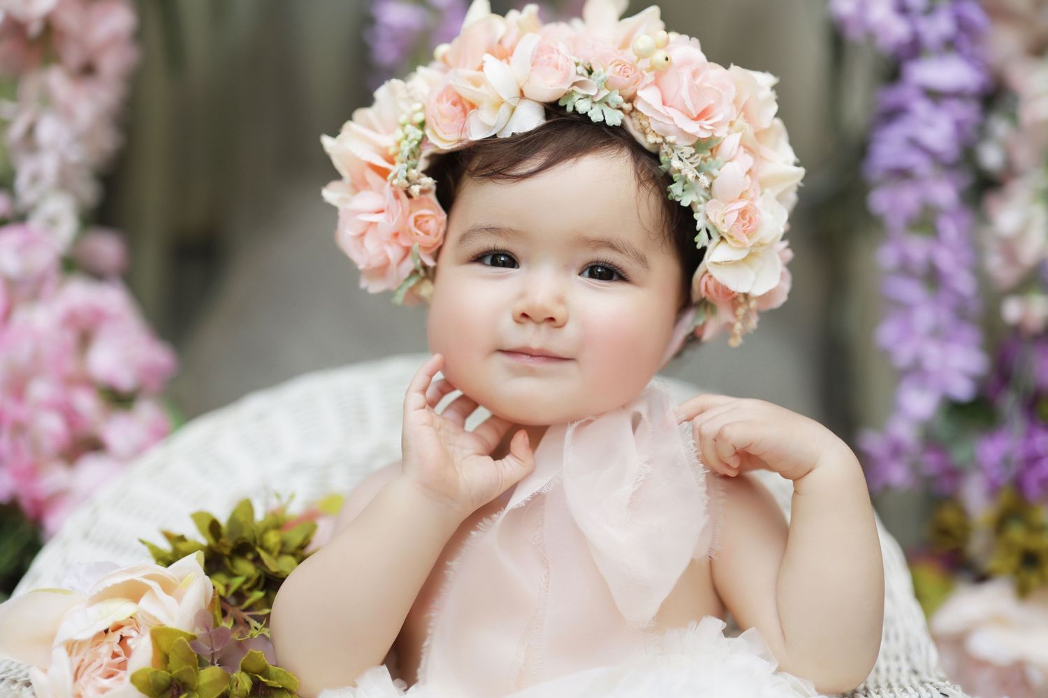 Adorable baby girl wearing pink floral crown surrounded by colorful flowers.