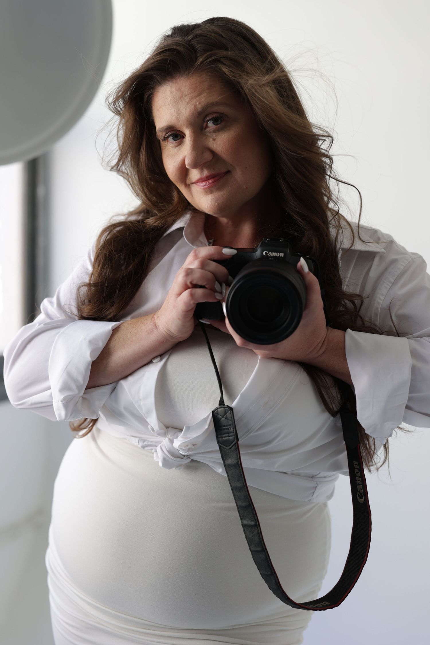 Pregnant woman in white shirt holding a Canon camera in a bright studio setting.