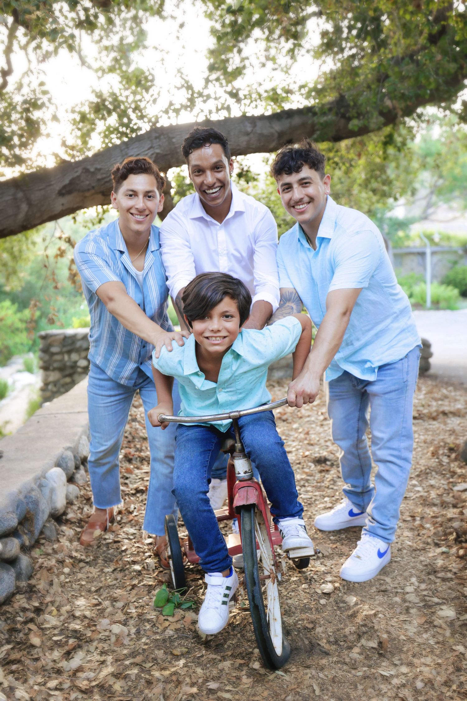 Family of four smiling outdoors as young boy rides red bicycle under a tree on a sunny day.