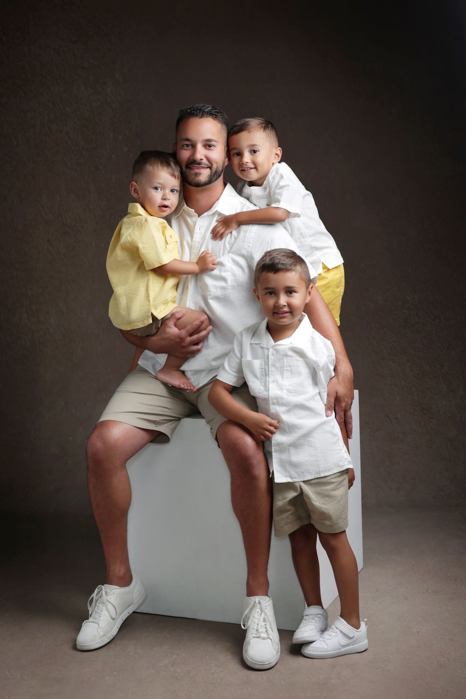 A smiling father poses with his three young sons in matching outfits for a professional studio family portrait.