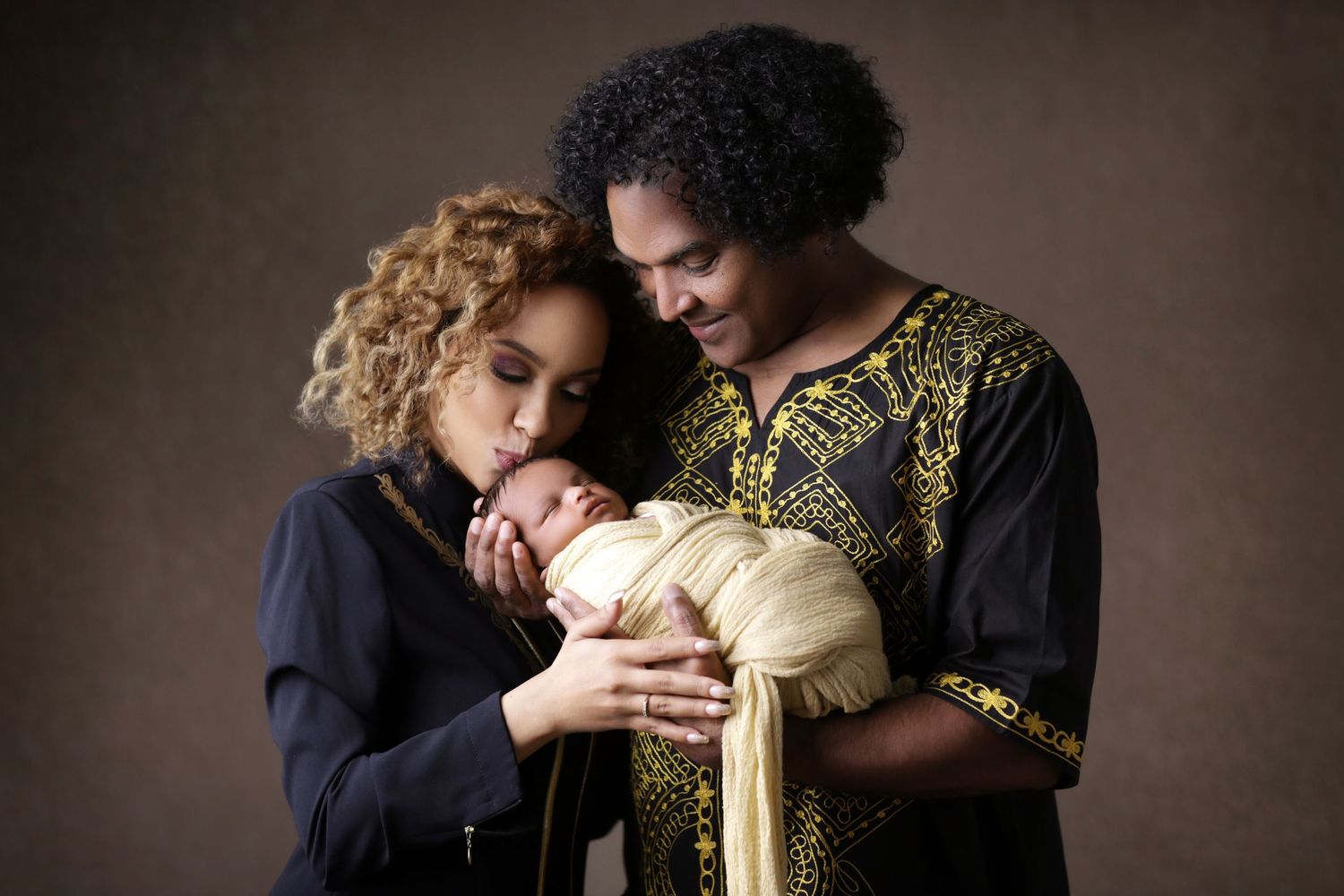 Two parents lovingly cradle their newborn baby wrapped in cream fabric against a warm brown studio backdrop.