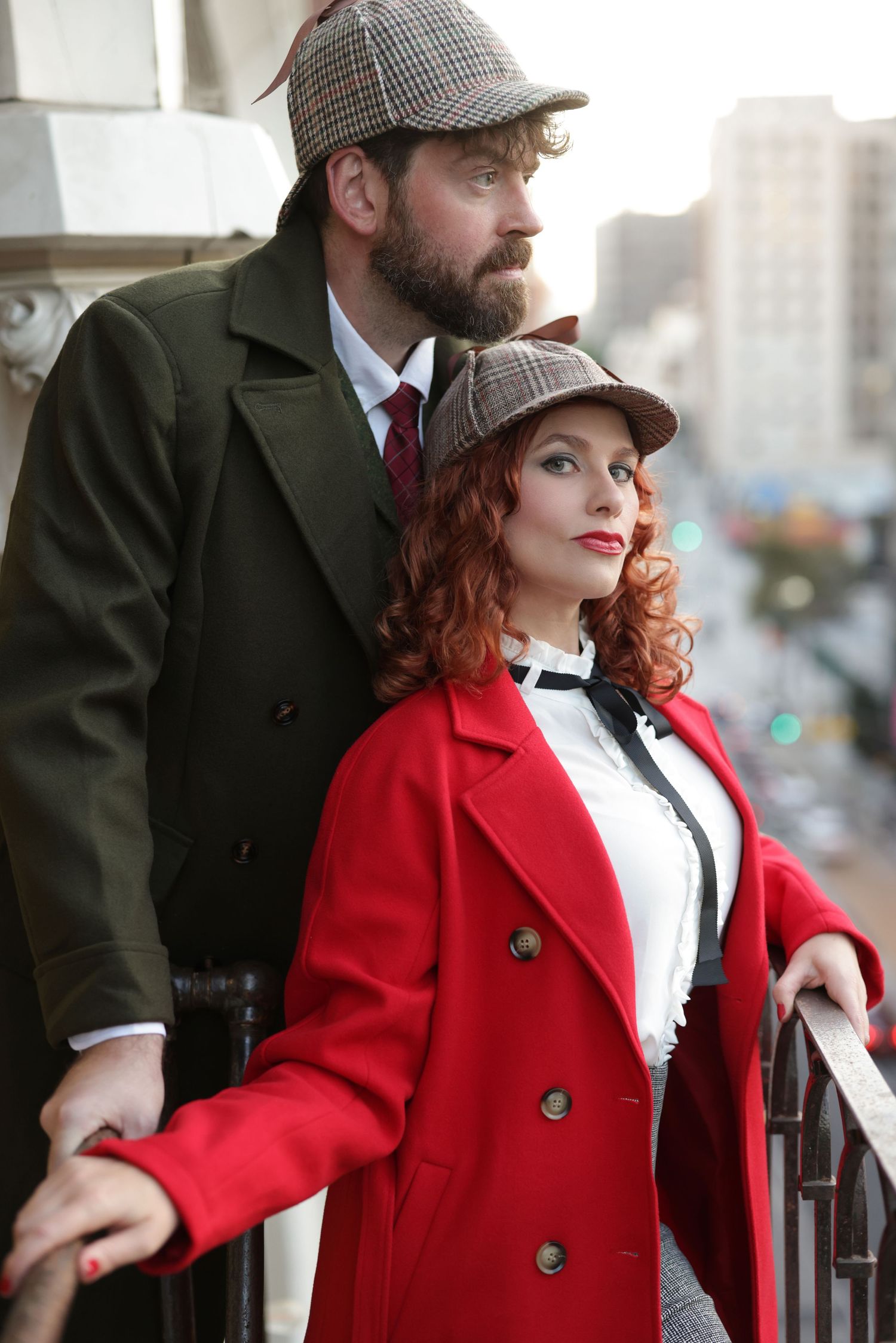 Elegant couple in vintage detective costumes on a balcony, man in tweed cap and woman in red coat.