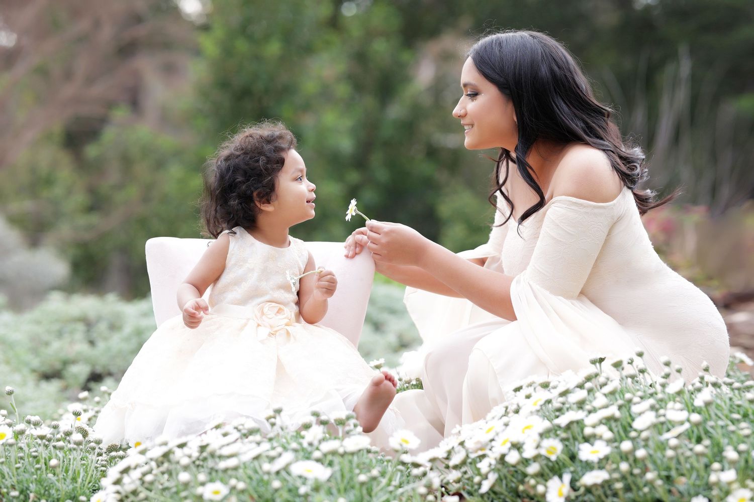 A mother and toddler daughter in matching white dresses share a tender moment among white flowers in a garden.