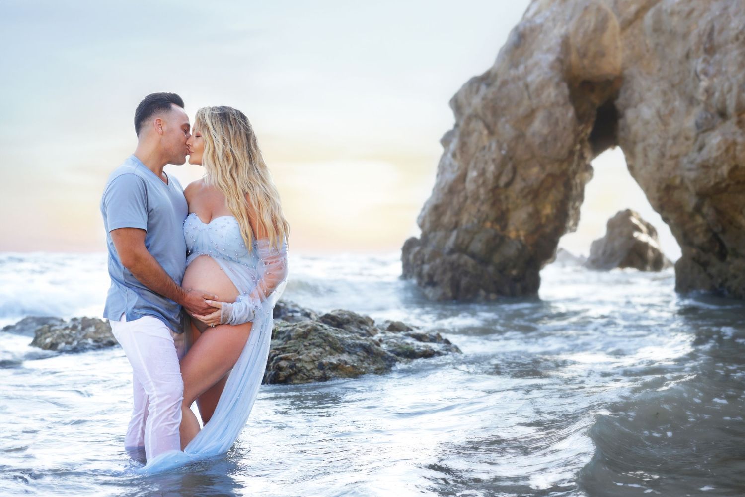 Couple sharing a kiss on a rocky beach during a maternity photoshoot, waves crashing around them at sunset.