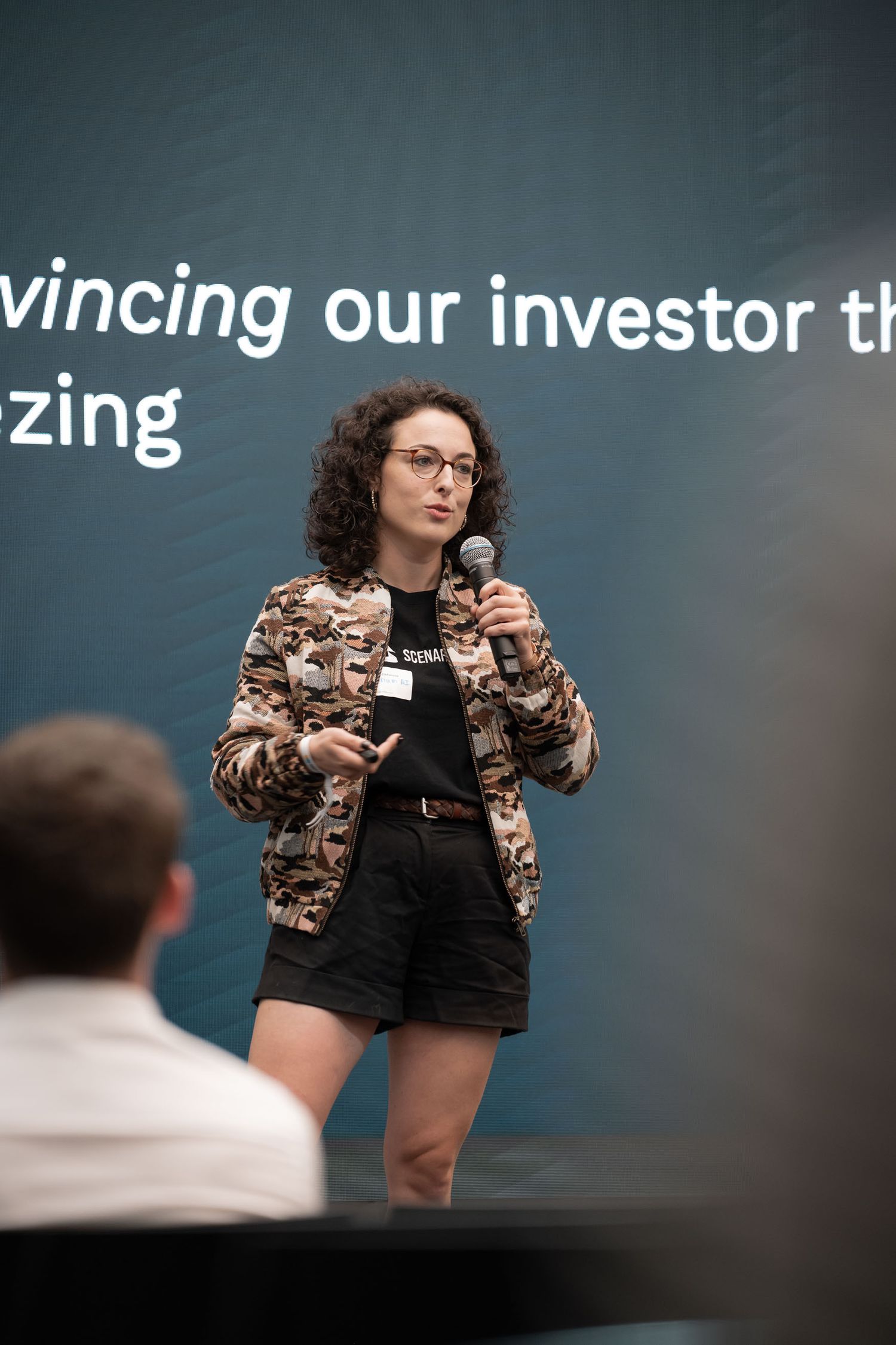 Professional speaker in animal print blazer and black outfit gestures while presenting at business conference.