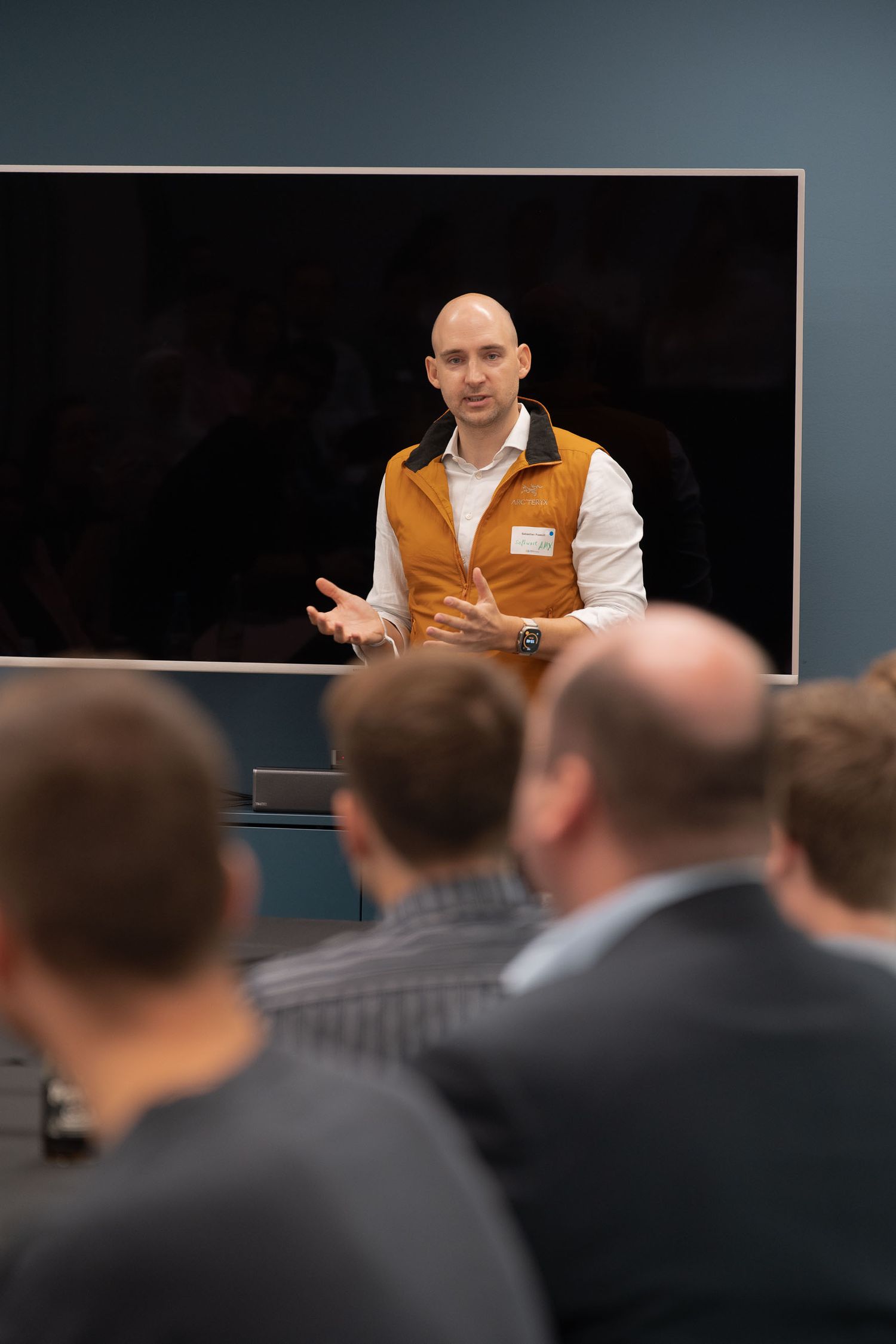 A presenter in an orange vest speaks to an audience during a business conference or training session.