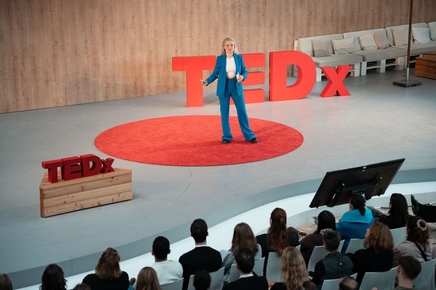 A speaker stands on a red circular stage with large TEDx letters during a presentation to an audience.
