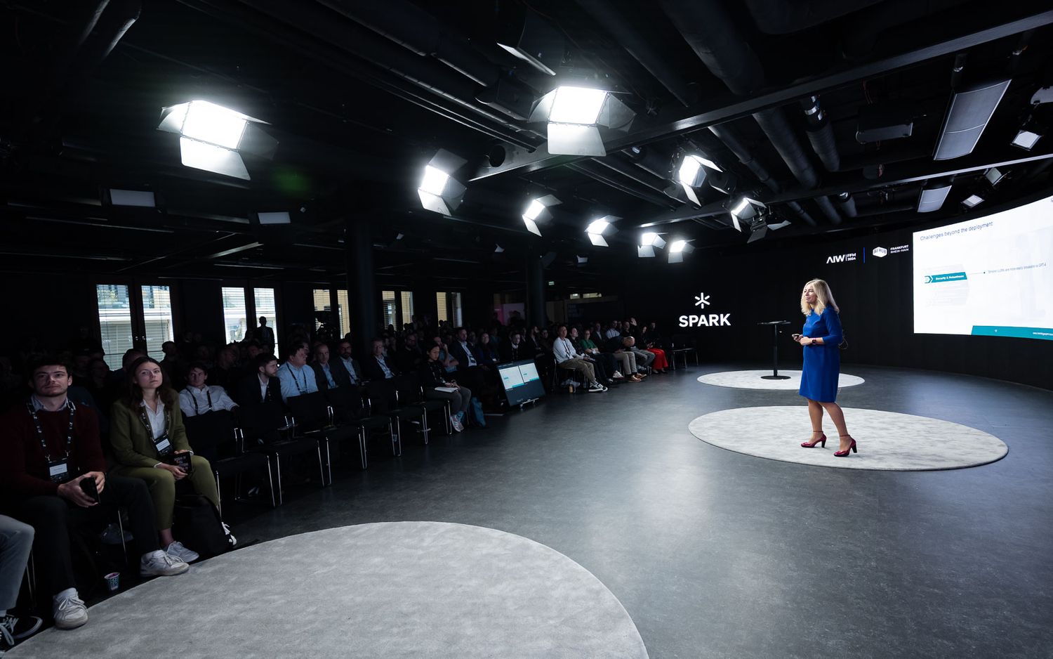 A dark fashion runway show with spotlights illuminating a circular stage as spectators watch from the sides.