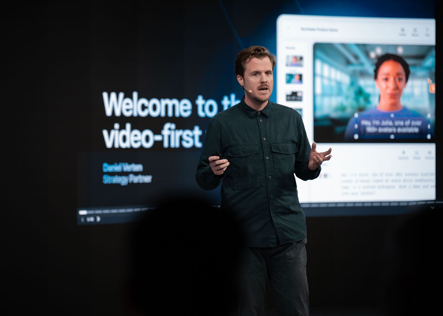 A presenter speaks on stage at a tech conference with a large screen displaying 'Welcome to video-first' behind them.