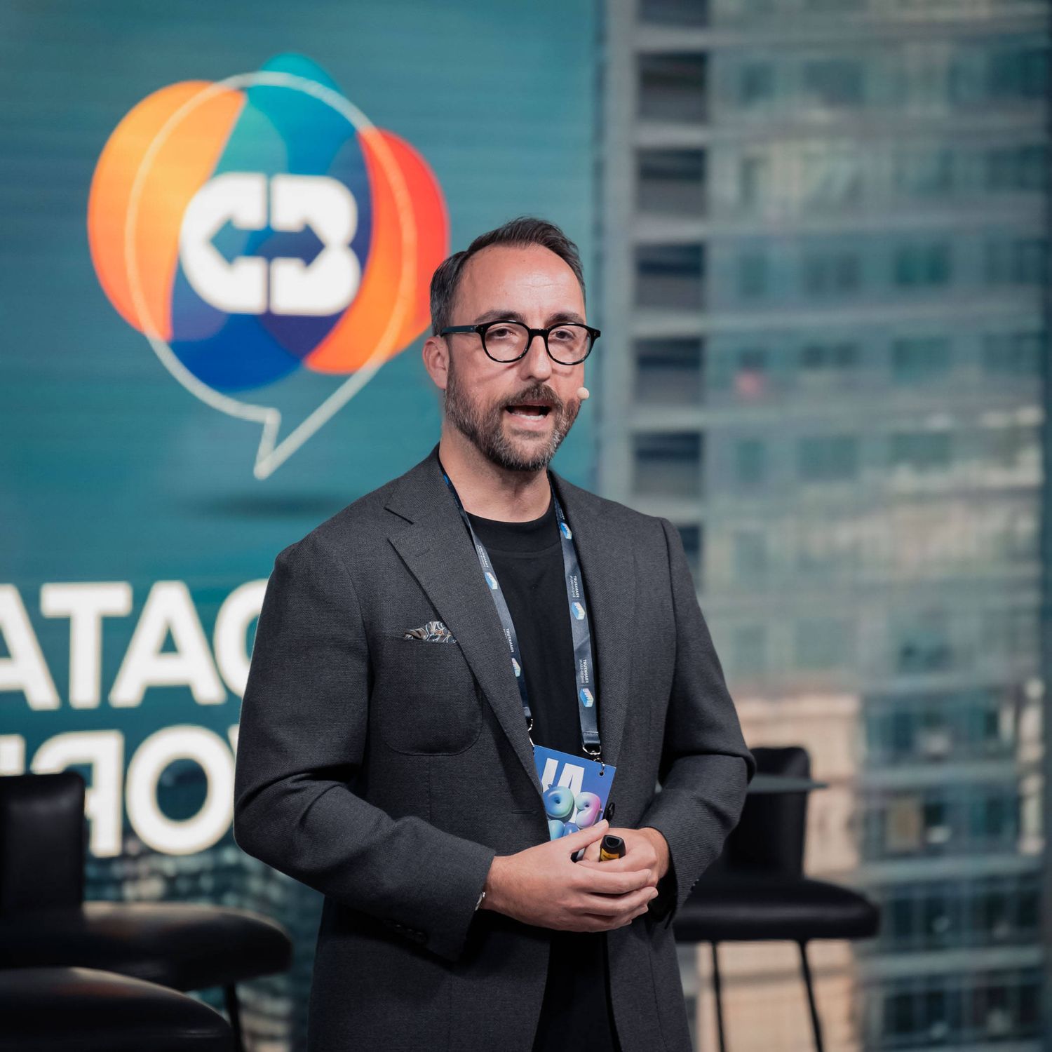 Speaker wearing glasses and dark attire presents on stage at a business conference with blue lighting.