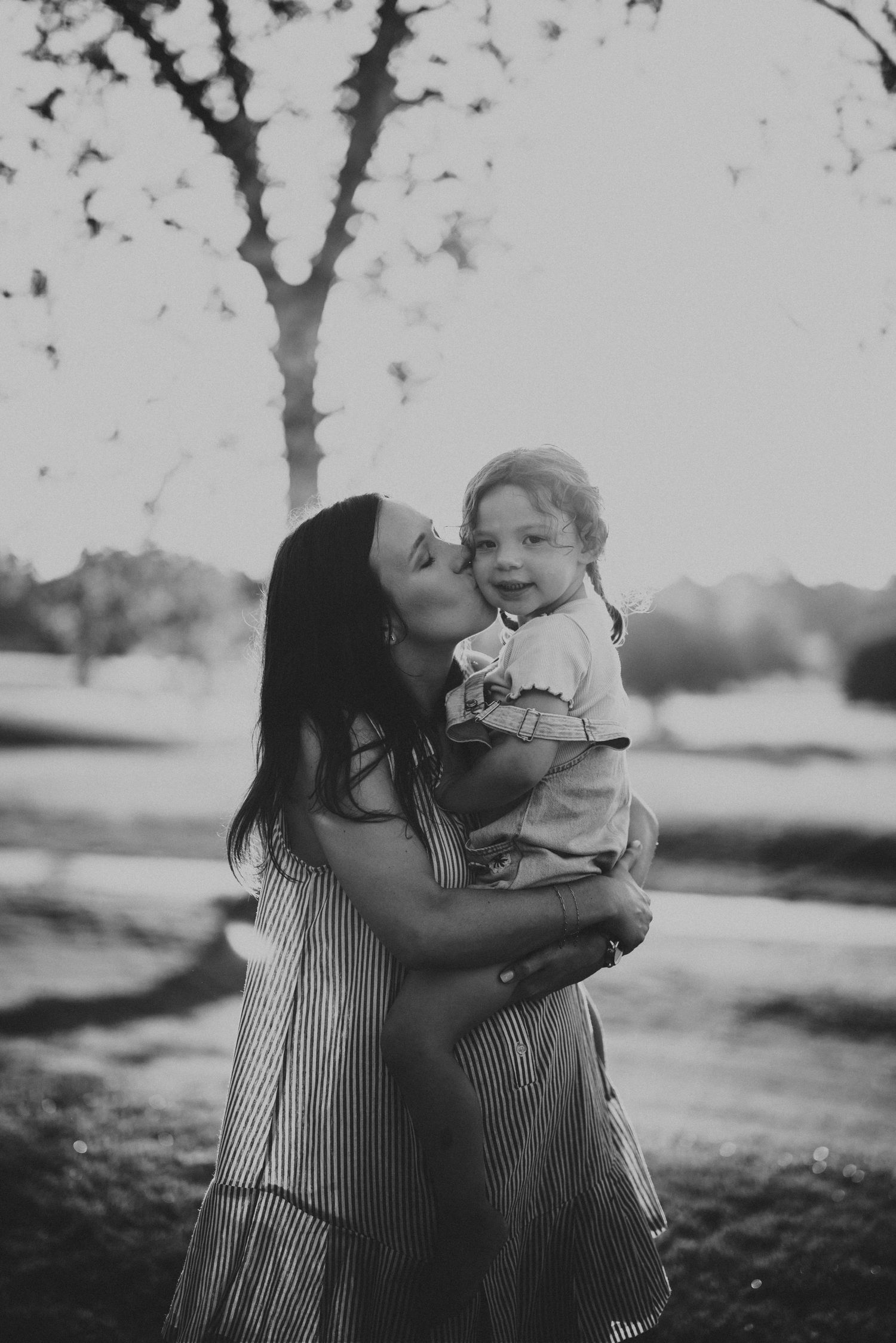 Black and white photo of a mother kissing her smiling toddler outdoors in a park setting.