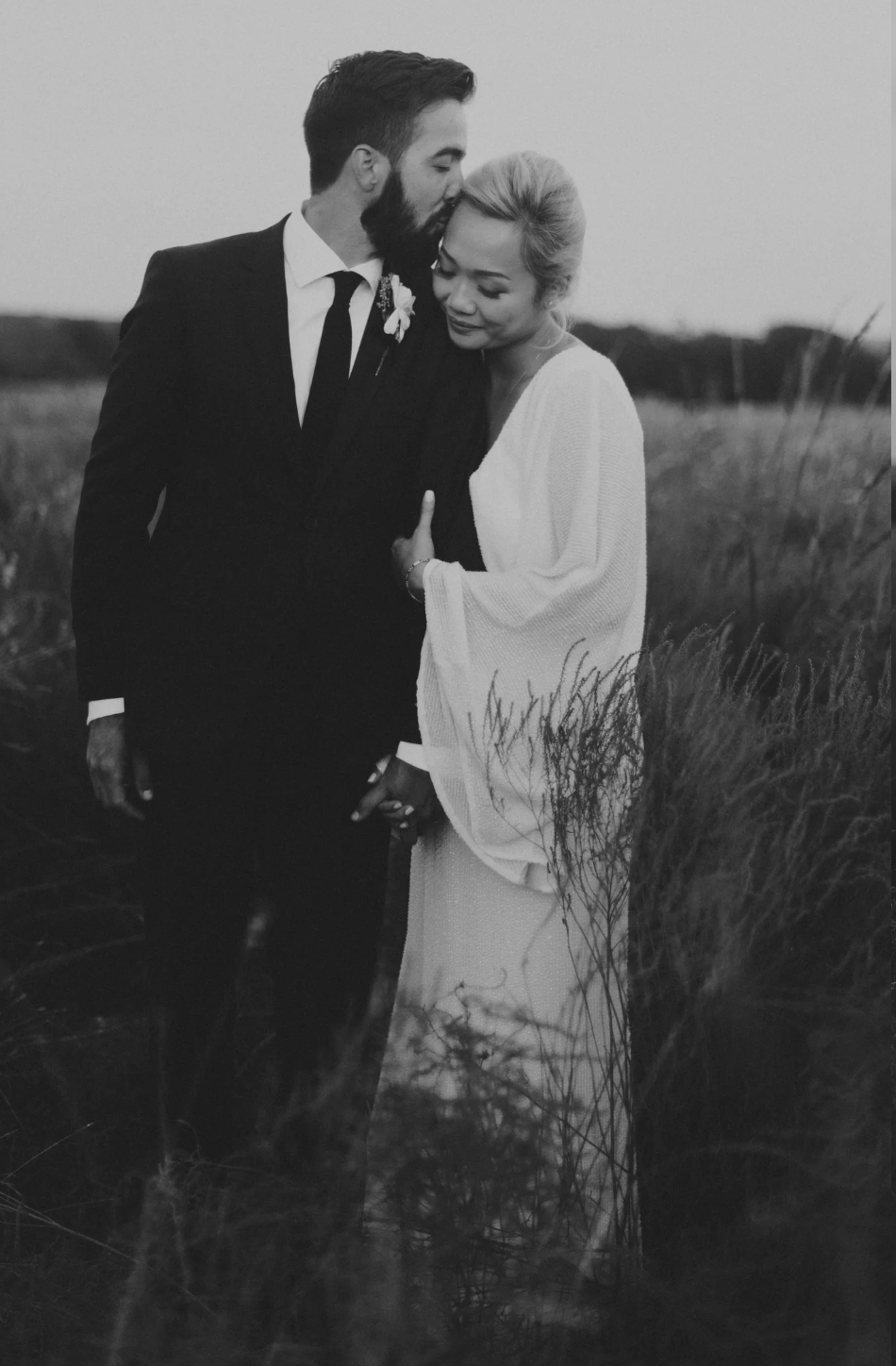 Black and white photo of groom kissing bride on cheek in a tall grass field at dusk.