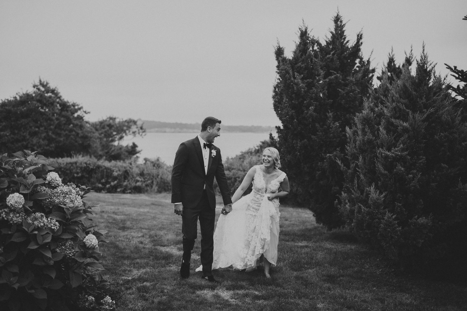 Black and white photo of bride and groom strolling hand in hand through lush garden near waterfront.