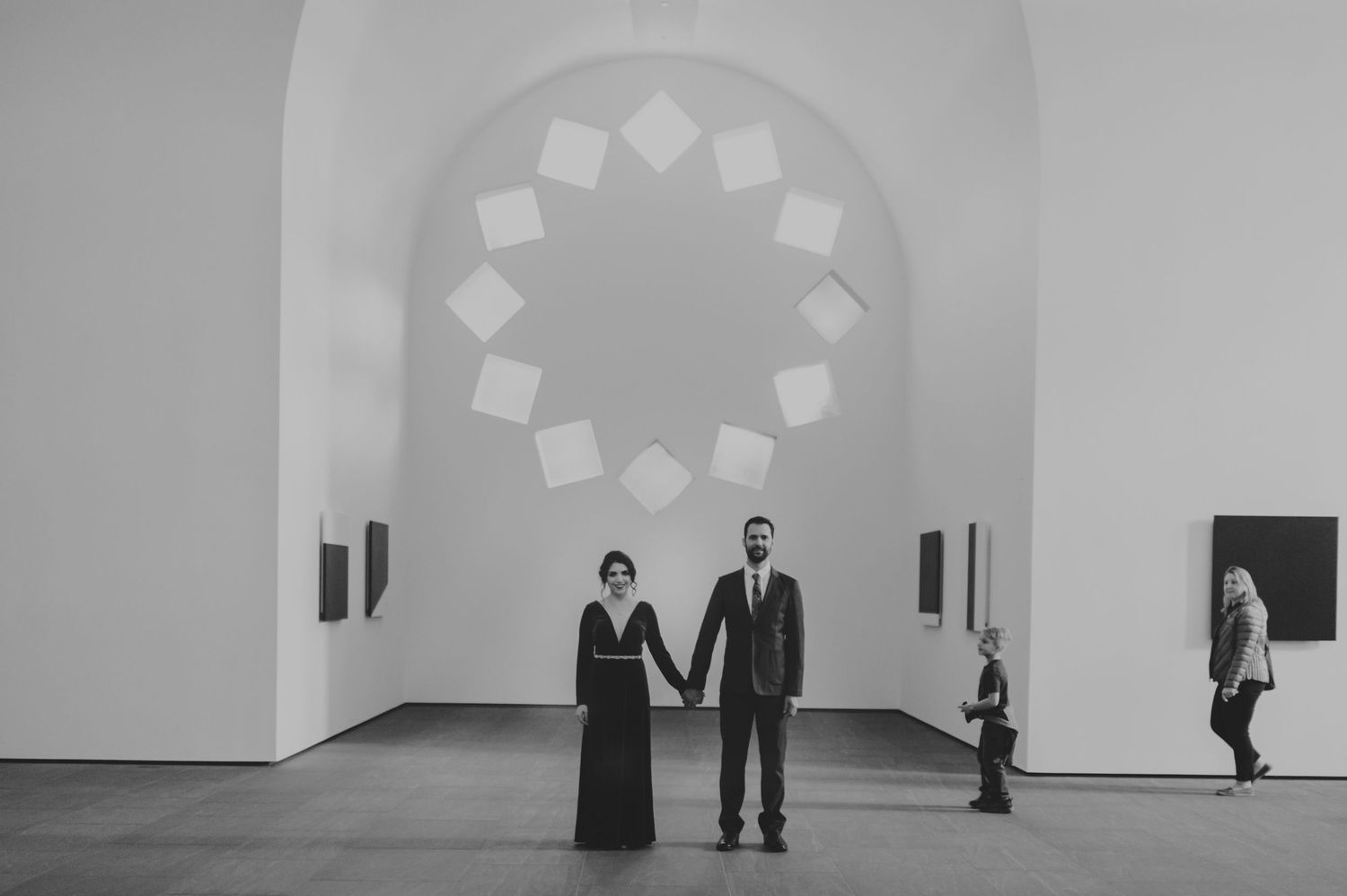 Couple holds hands in black and white photo inside art gallery with circular window.