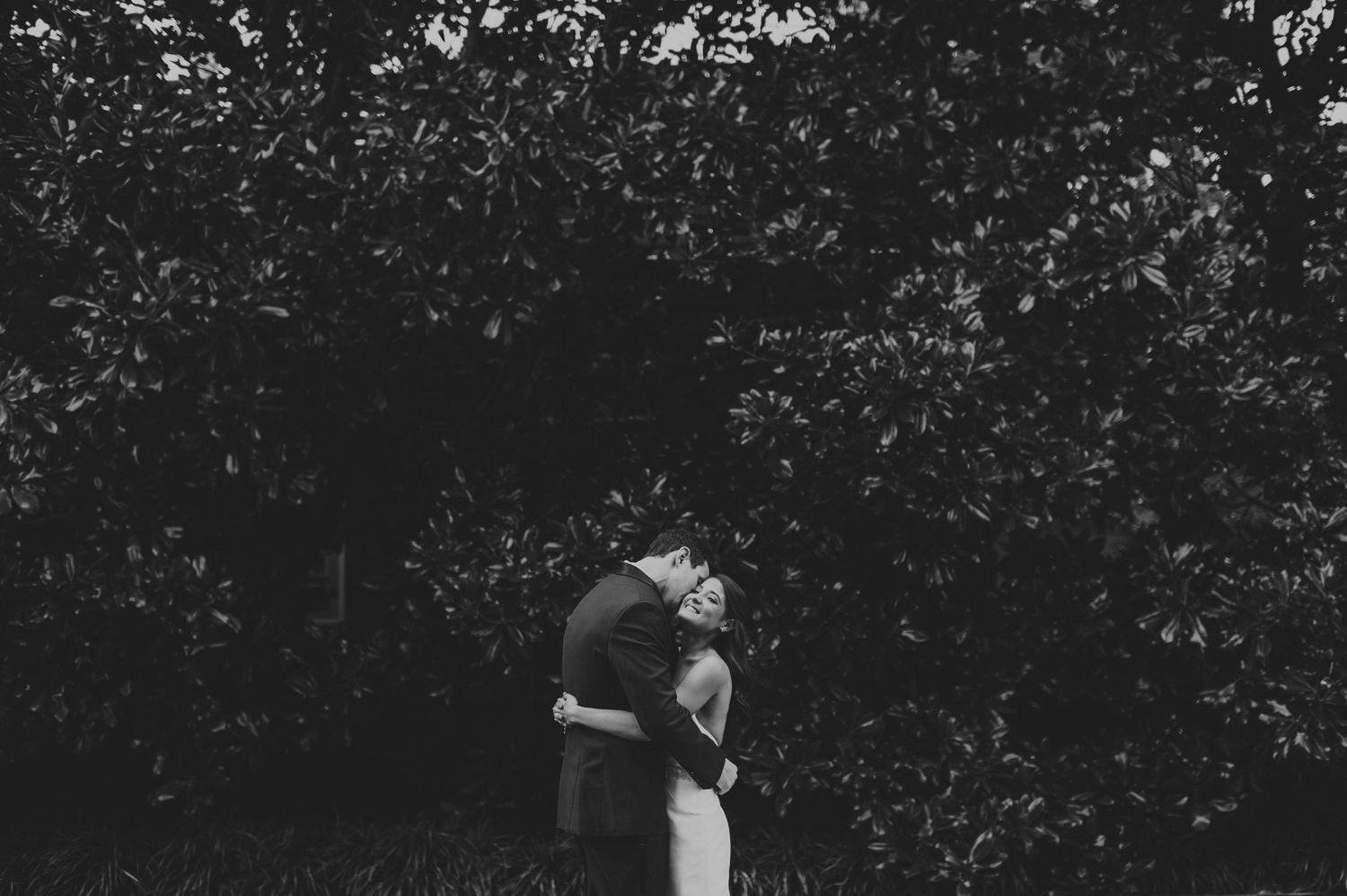 Black and white photo of a bride and groom embracing under lush green trees outdoors.