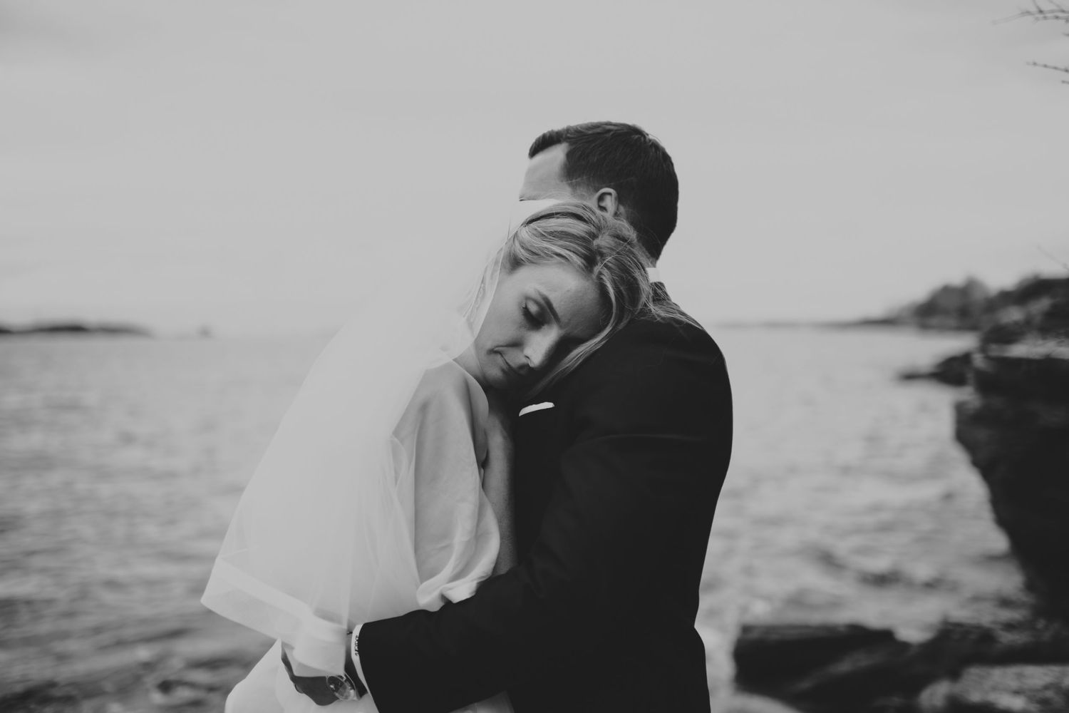 Bride rests her head on grooms chest in a tender black and white wedding embrace by the sea.