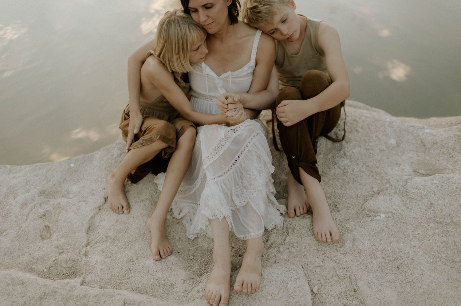 A mother in a white dress sits on a rock embraced by her two blonde children near water.