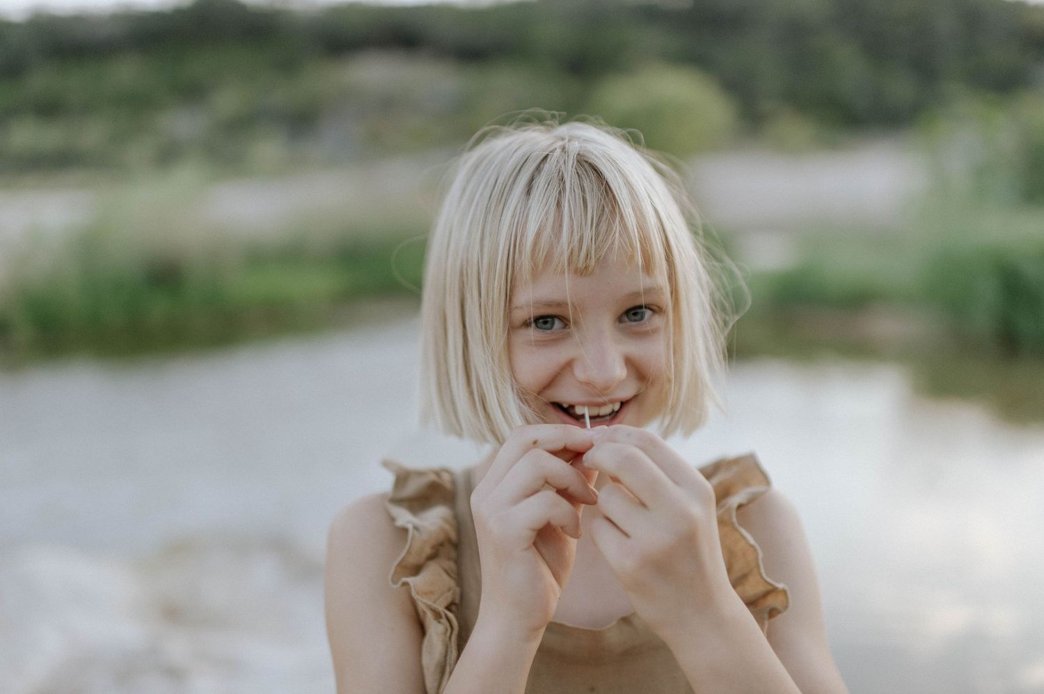 Happy blonde girl with bangs smiling playfully near a lake outdoors.