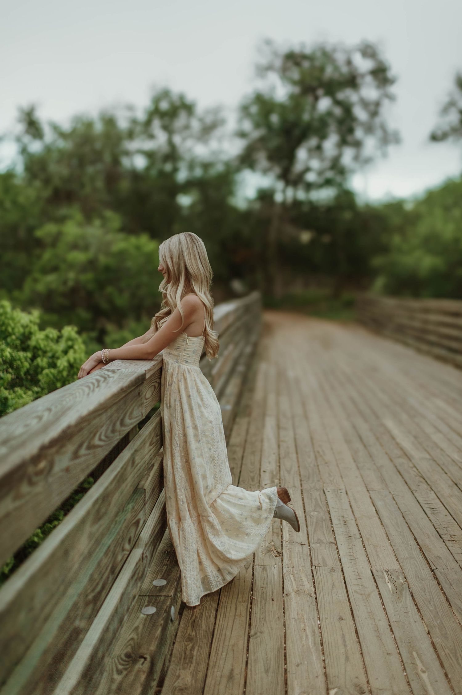 Blonde woman in a floral maxi dress leaning on a wooden bridge railing surrounded by greenery.