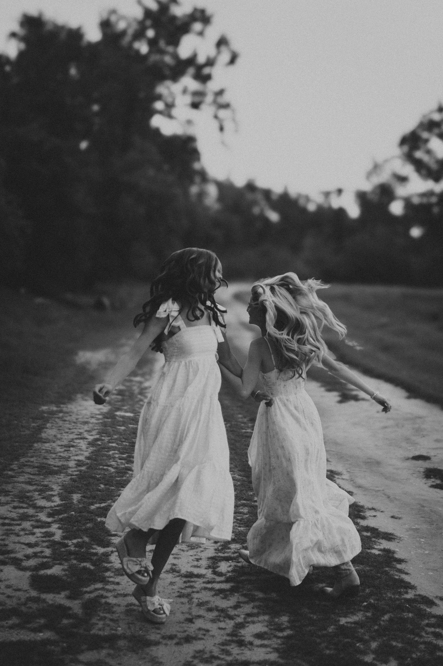 Two girls in white dresses holding hands and running down a rustic path in a dreamy black and white photo.