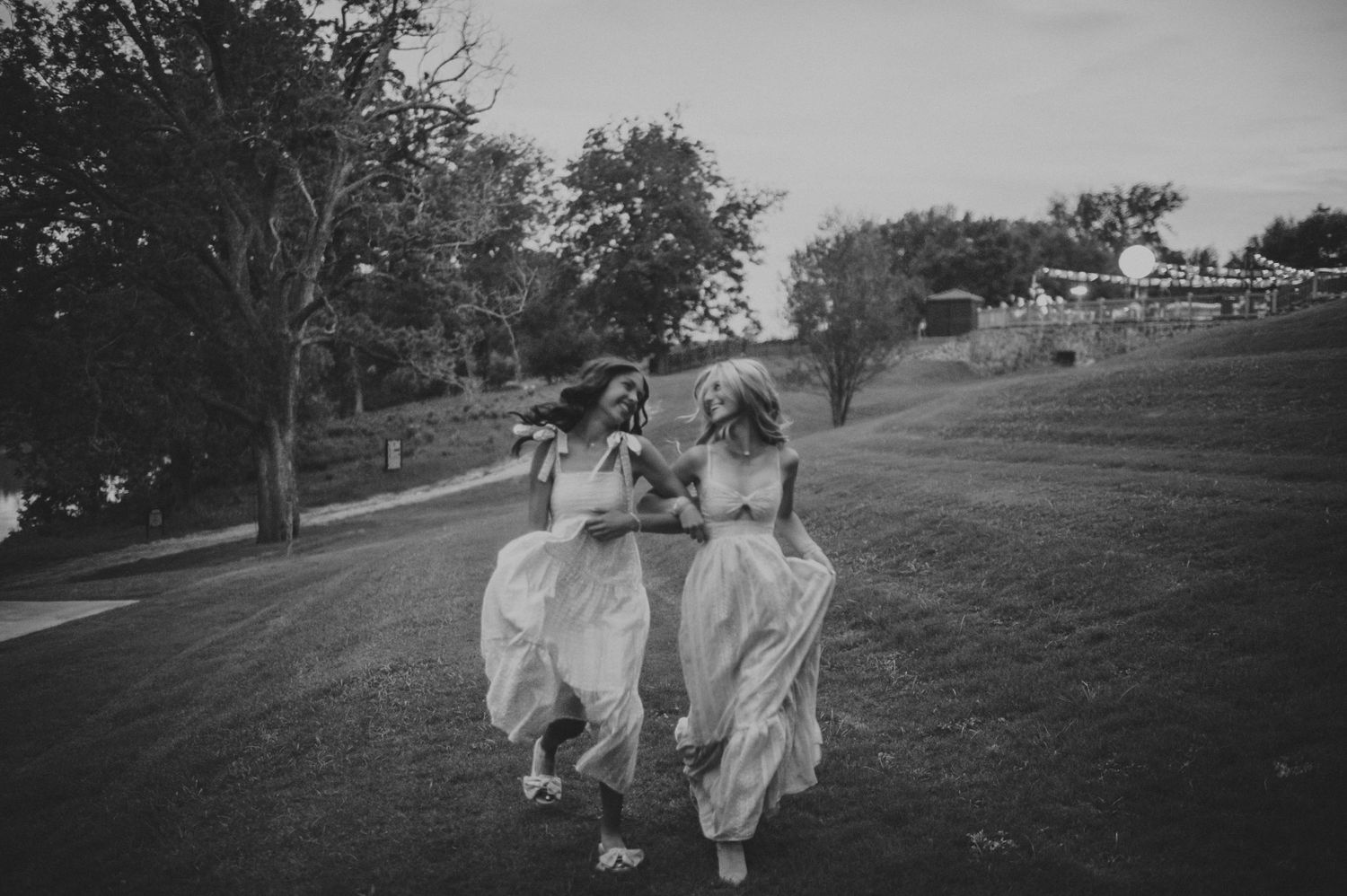 Two young women in white dresses walk and laugh together across a grassy hill in black and white.