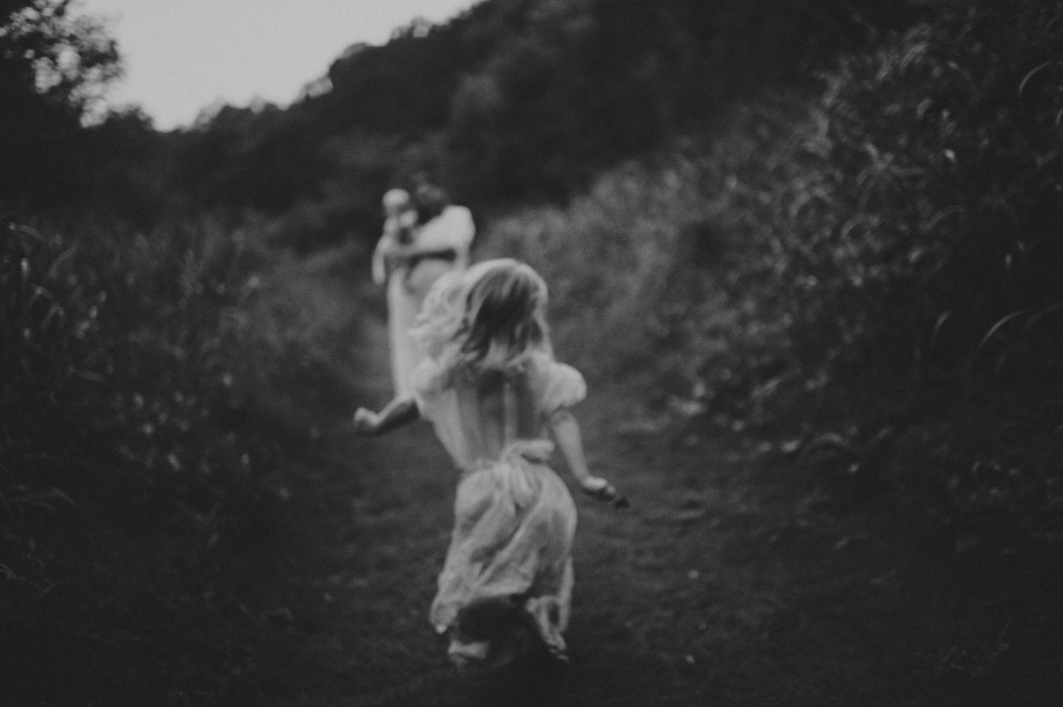 Two young girls in white dresses running down a nature path in black and white.