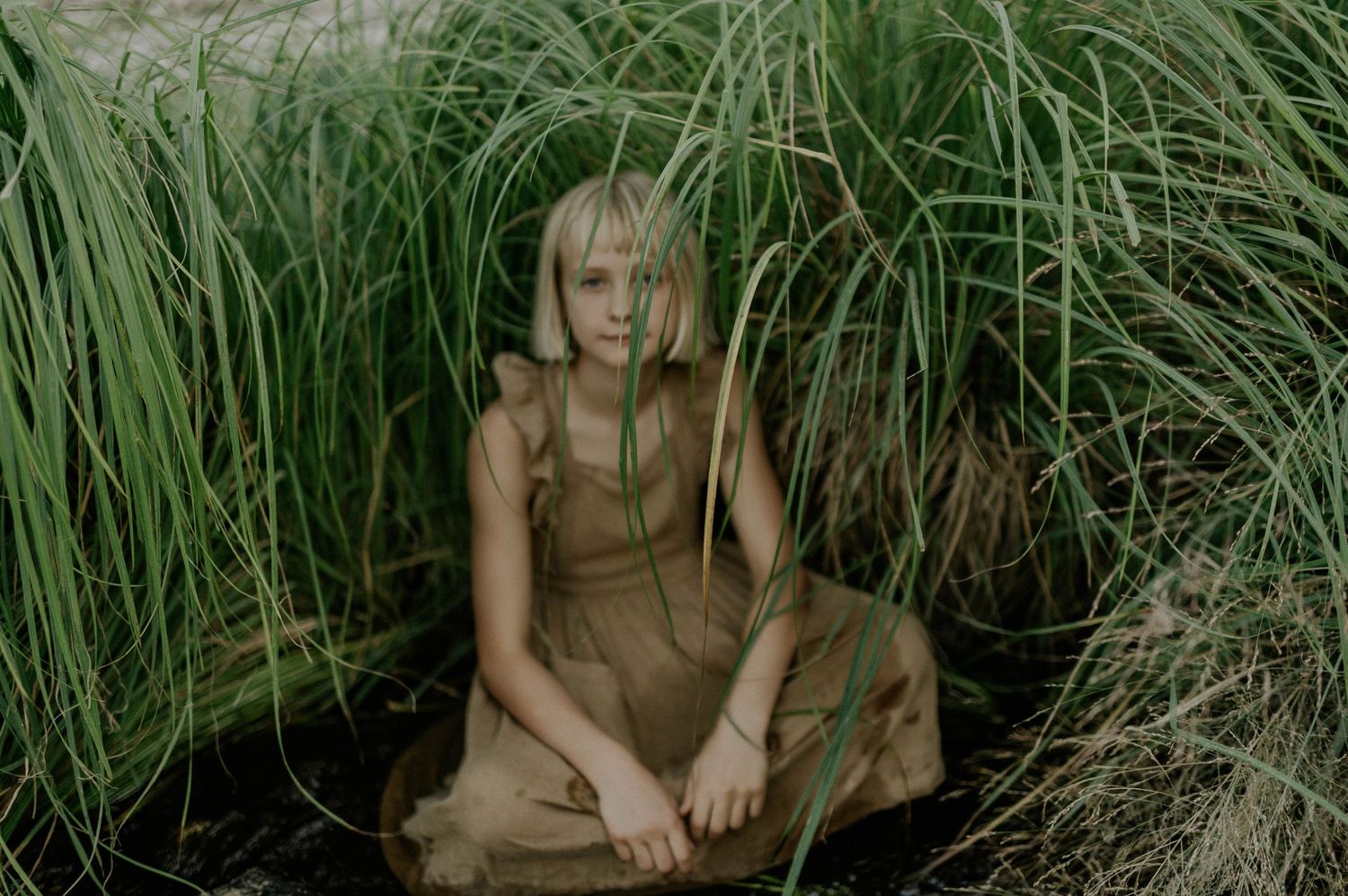 A young blonde girl in a tan dress sits peacefully surrounded by tall green grass.
