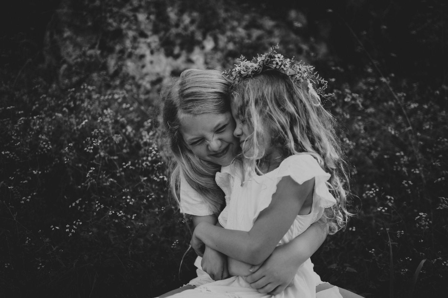 A smiling woman hugging a little girl wearing a floral crown in a lush garden setting.
