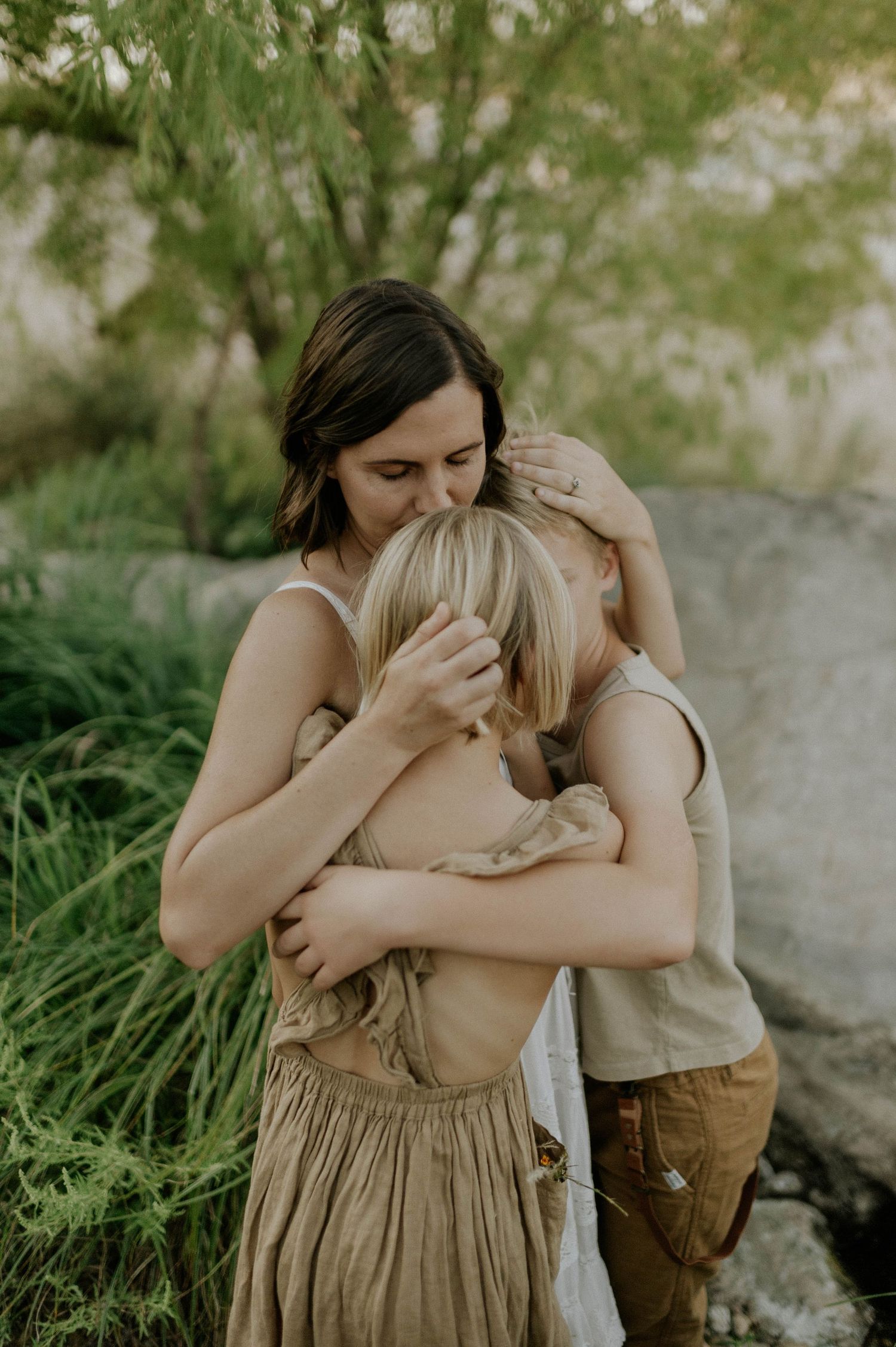 Two women embrace a child tenderly outdoors, surrounded by lush greenery in a warm, natural setting.