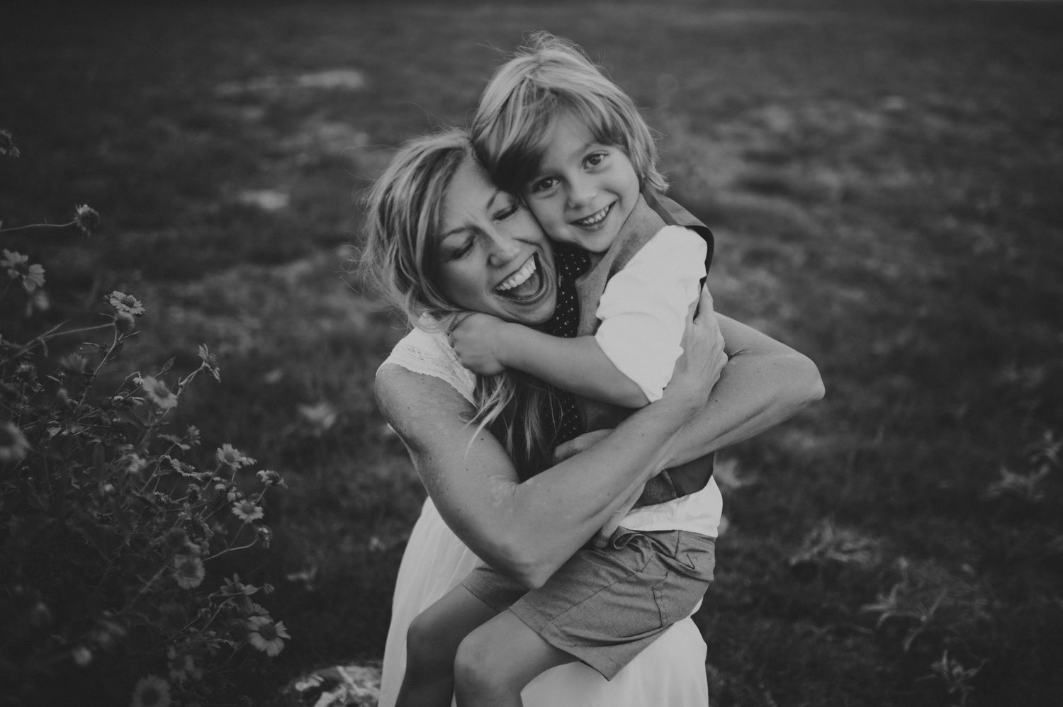 Black and white photo of a laughing woman hugging a young blonde child outdoors on a grassy background.