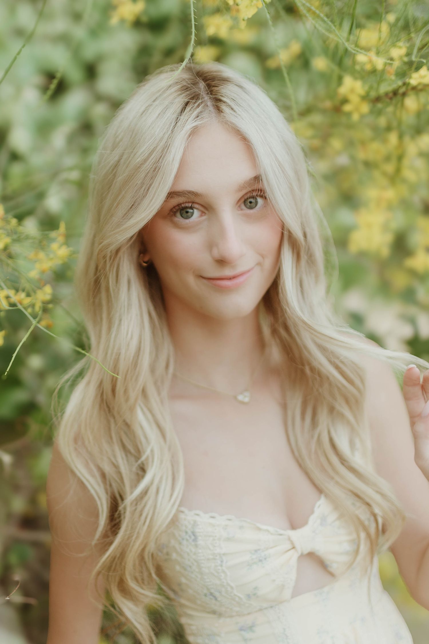Blonde teenage girl with long wavy hair in a cream strapless dress posing among yellow flowering trees.