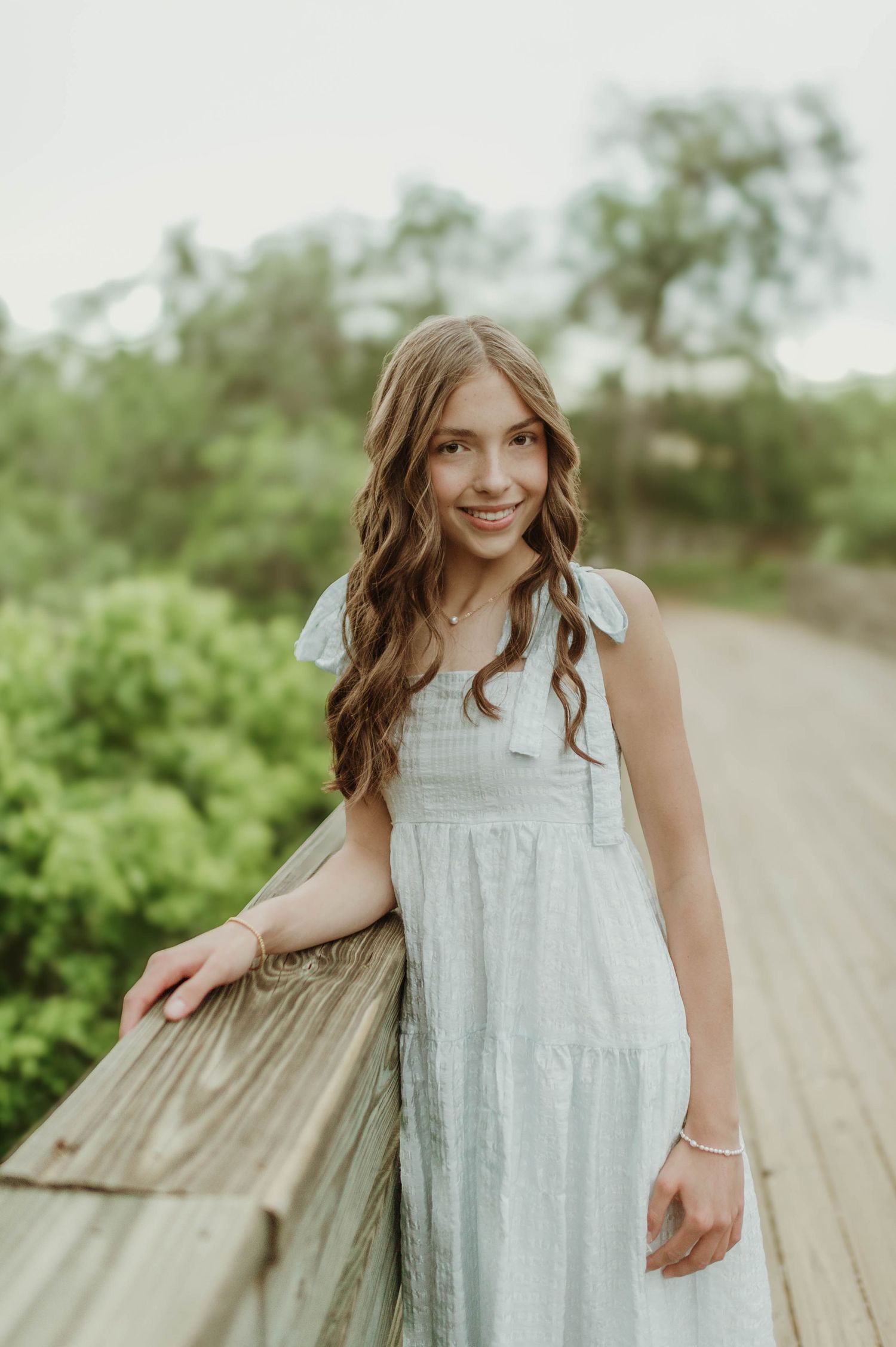 Smiling young girl with wavy brown hair in a light blue sundress on a wooden boardwalk.