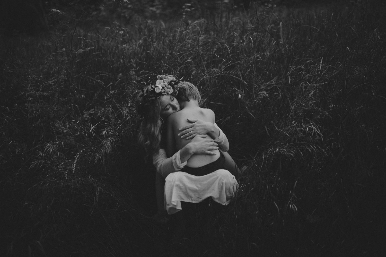 Black and white photo of a woman with floral crown tenderly holding a child in a meadow.