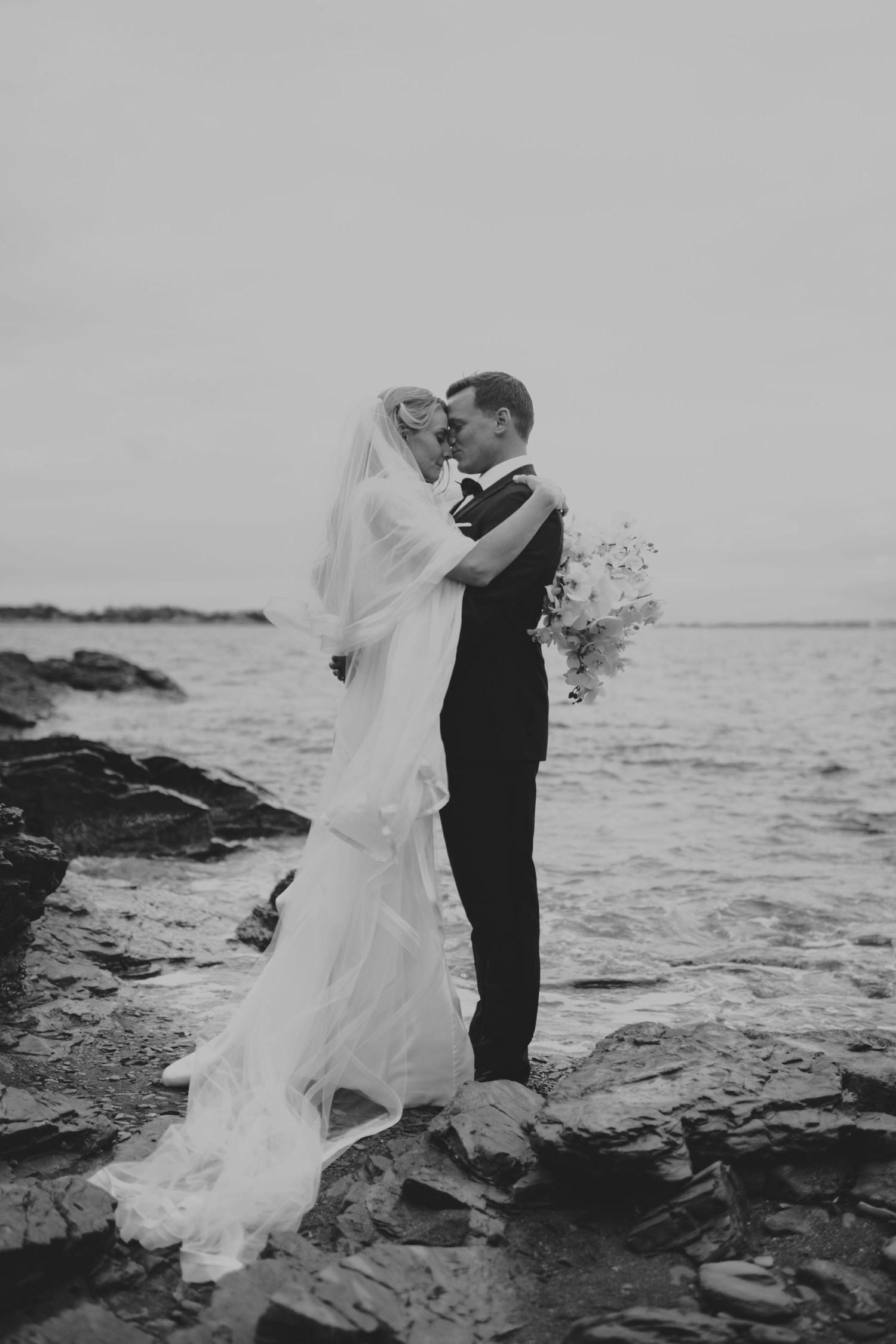 Bride and groom kiss while standing on rocky shoreline with ocean waves in black and white photo.