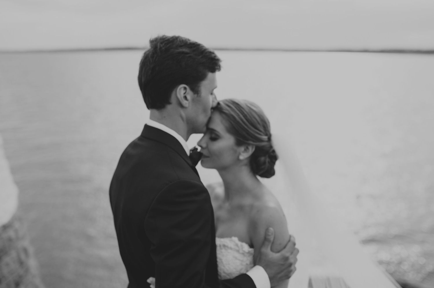 Bride and groom share a tender kiss by the calm ocean waters in a black and white portrait.