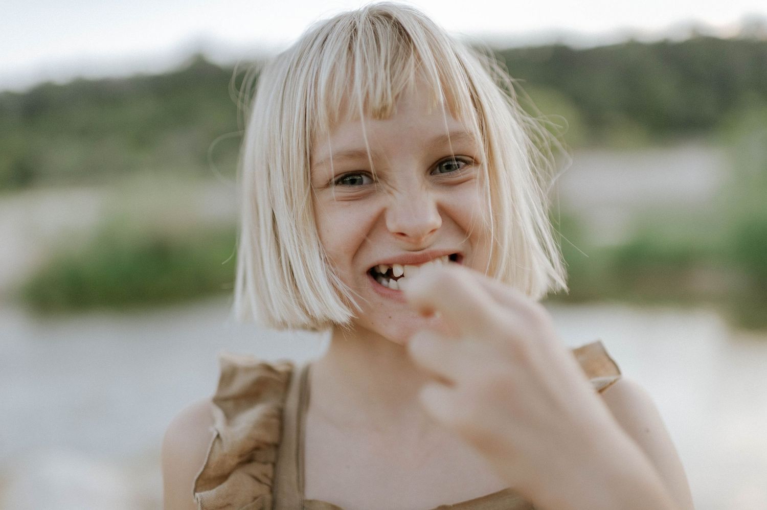 Smiling blonde girl with bangs pointing at her teeth outdoors near water.
