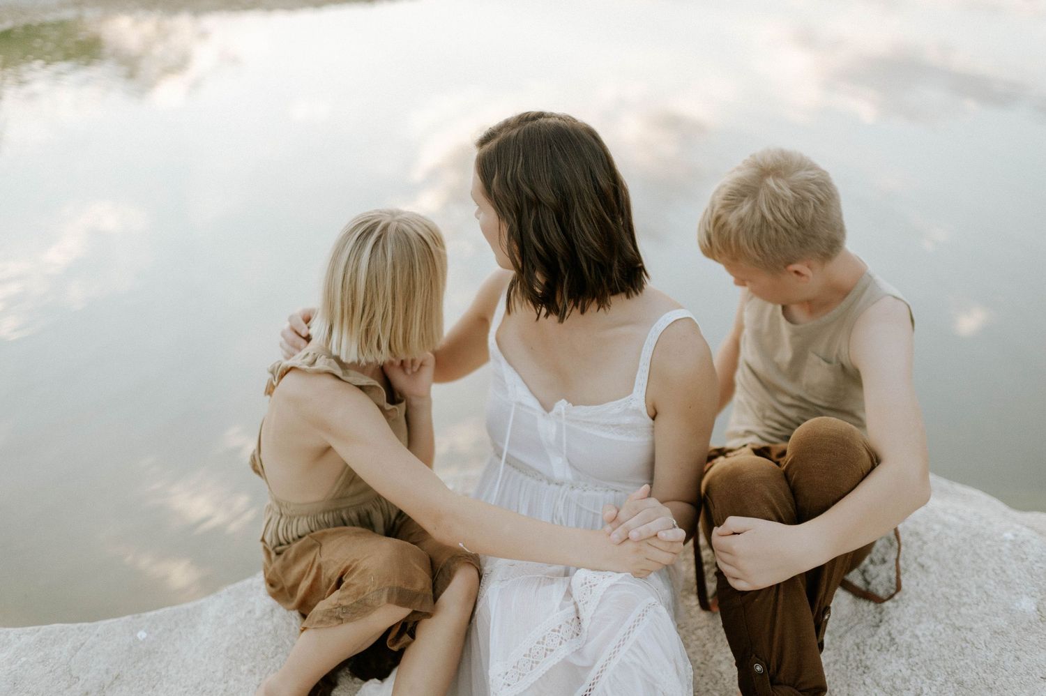 A mother in a white dress sits on a rock with her two children overlooking a peaceful lake.