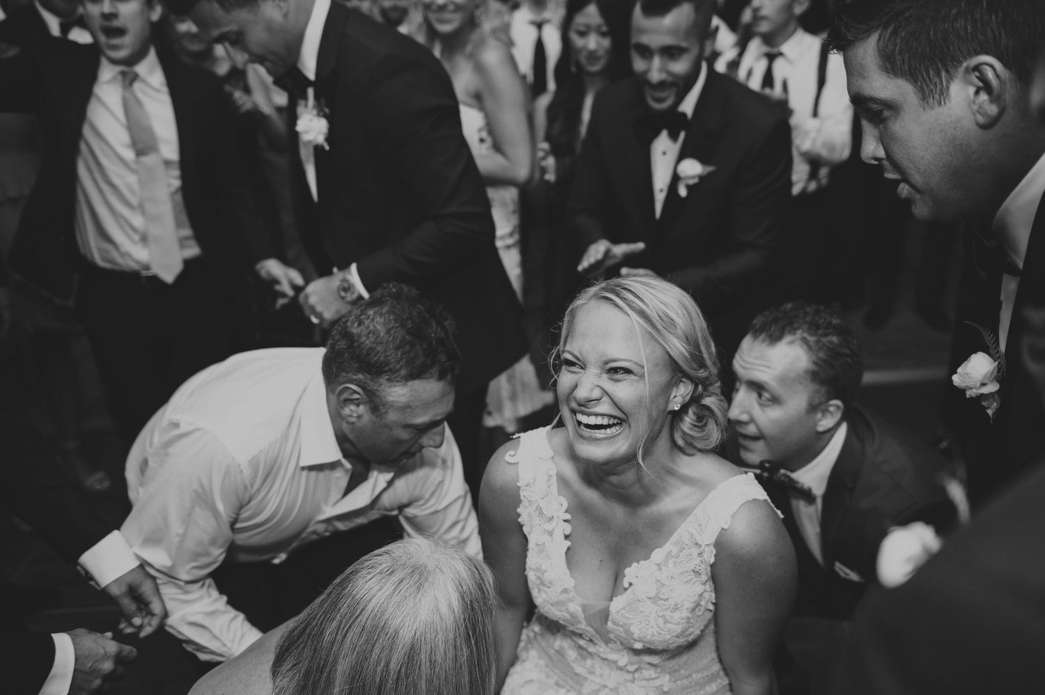Black and white photo of laughing bride surrounded by joyful wedding guests on the dance floor.