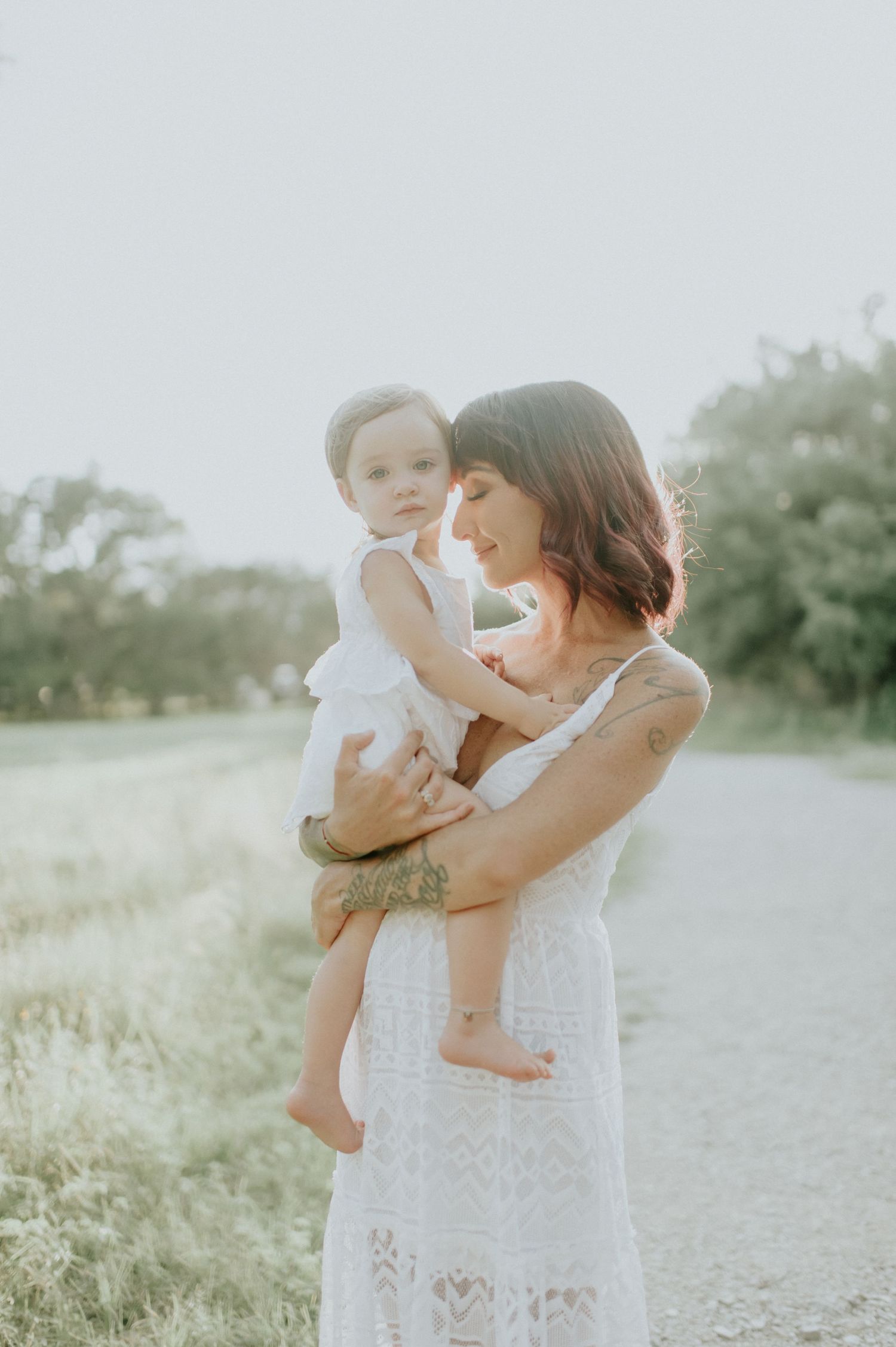 A mother in a white lace dress holds her baby outdoors on a sunlit path surrounded by greenery.