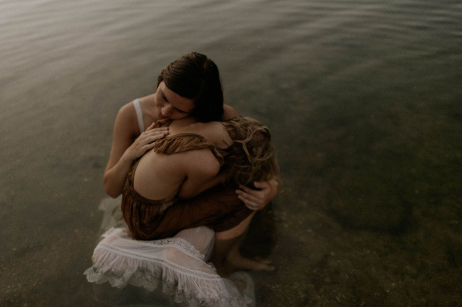 Two women share a tender embrace while sitting in calm shallow water wearing flowing dresses.