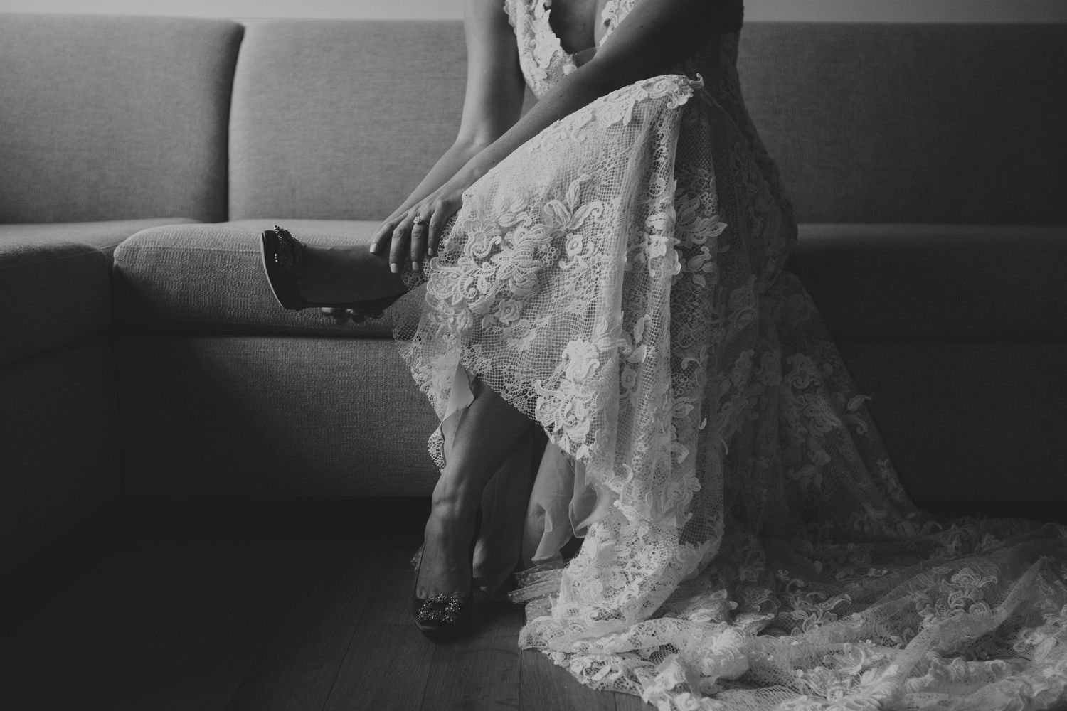 Black and white photo of a bride in lace wedding gown sitting on a sofa.