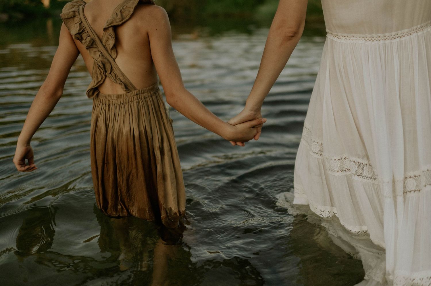 A young girl in a golden dress holds hands with a woman in white wading through water.
