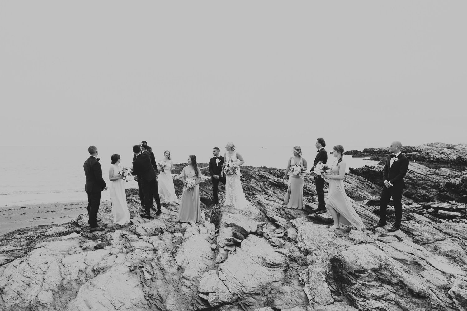 Black and white photo of wedding party gathered on rocky seaside cliffs.