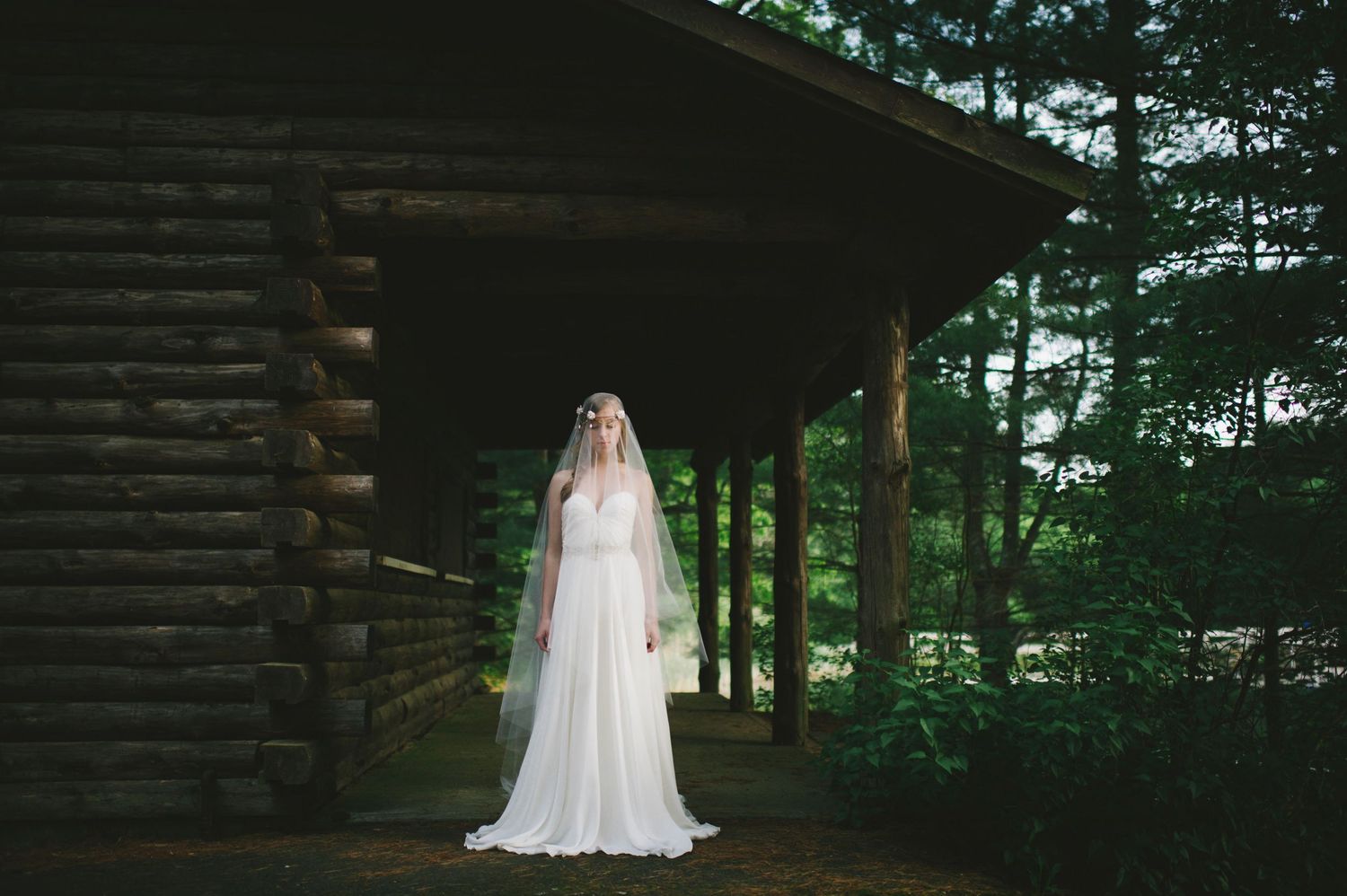 Bride in white gown with long veil standing before rustic log cabin surrounded by trees.