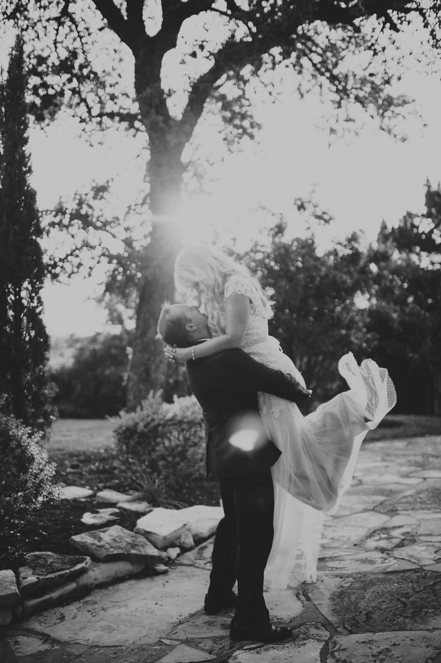 Groom lifts bride in a romantic embrace outdoors under a sunlit tree.
