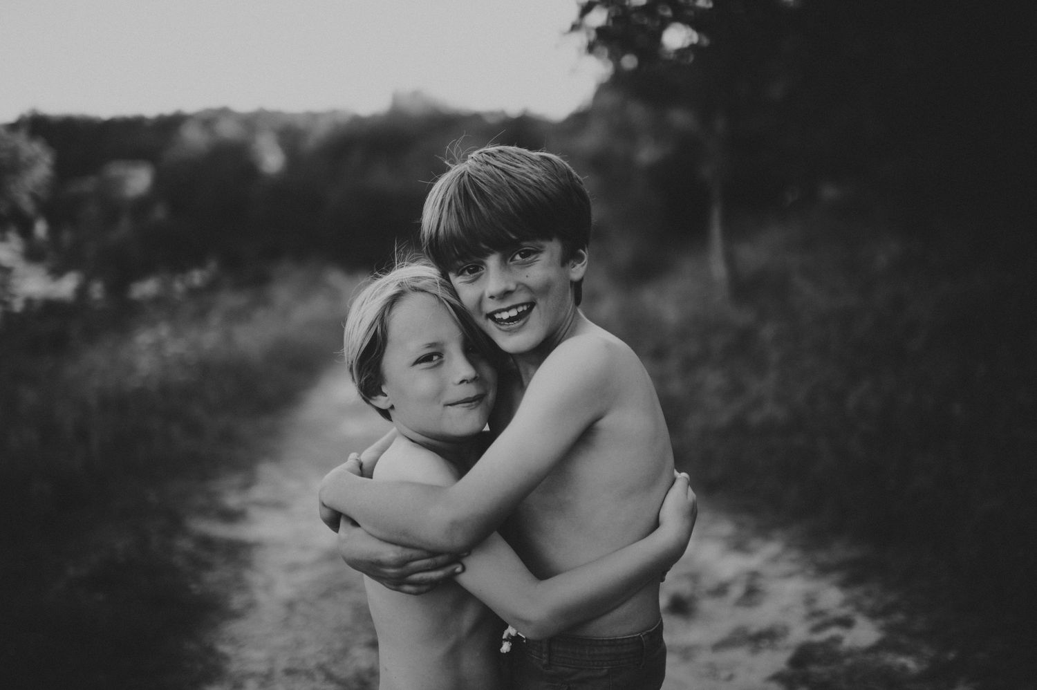 Two shirtless children hugging on a dirt path in a black and white outdoor portrait photo.