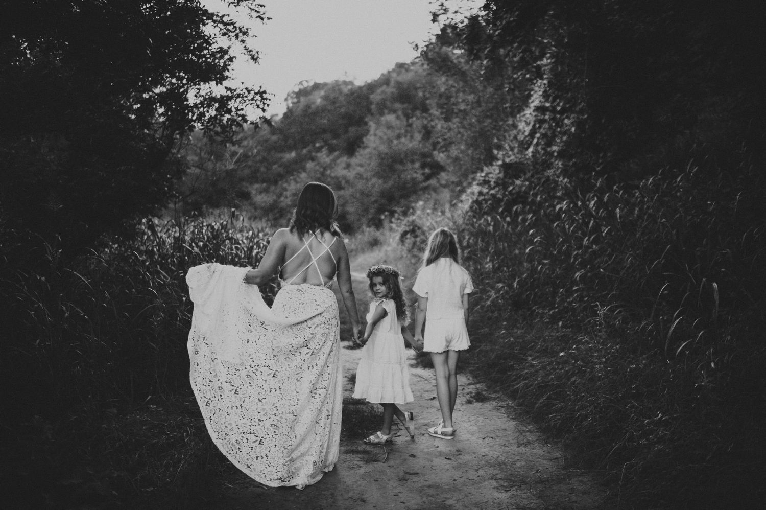 Black and white photo of women and girls in white dresses walking along a tree-lined path.