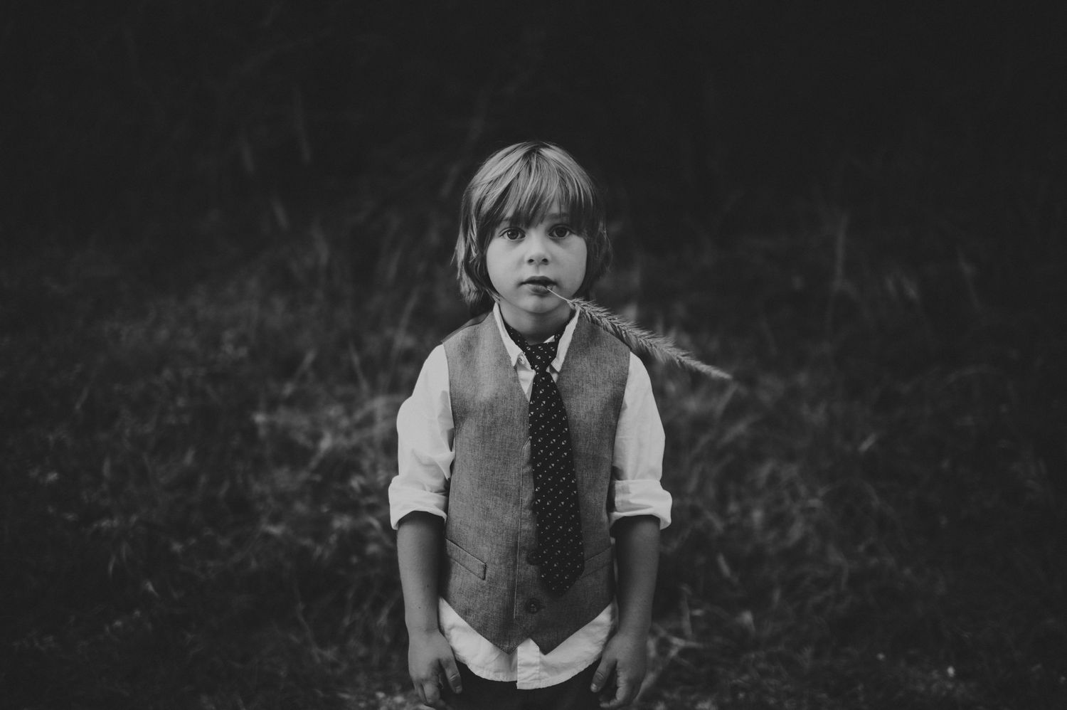 Black and white portrait of a young boy wearing a vest and tie standing outdoors in a field.